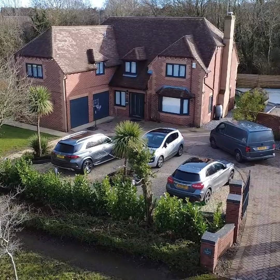 Drone view of a large detached house with a wide driveway, palms and cars parked
