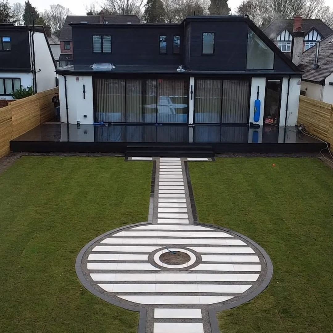A modern black and white house with a striped paving path leading to a large circular paved feature in a striped lawn
